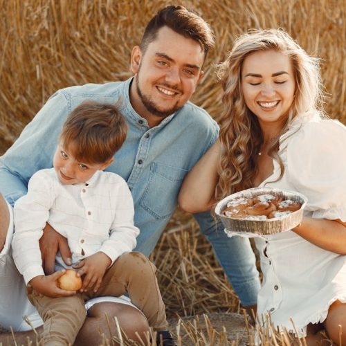 Smiling family of three enjoying a sunny day picnic with pastries on a hay field.