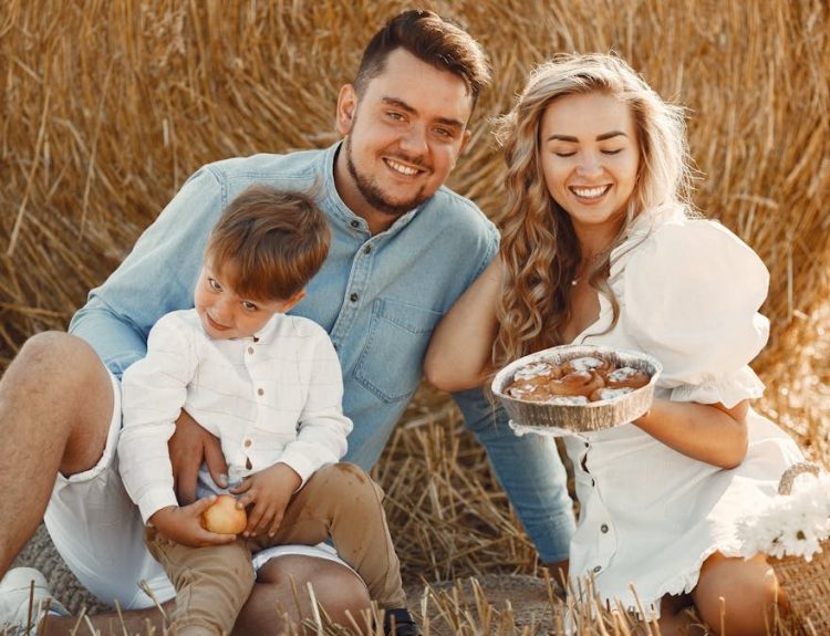 Smiling family of three enjoying a sunny day picnic with pastries on a hay field.