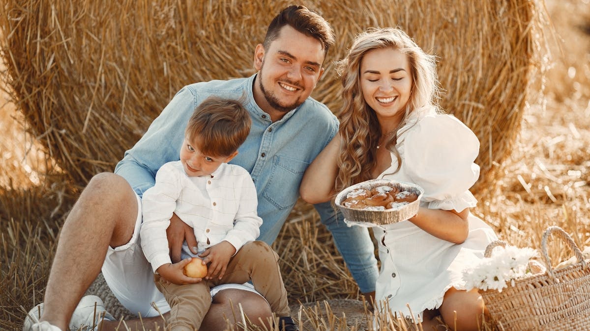 Smiling family of three enjoying a sunny day picnic with pastries on a hay field.